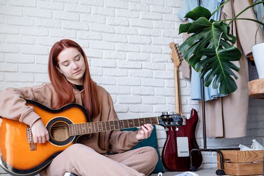 young caucasian red-haired woman sitting on rug and playing acoustic guitar at home