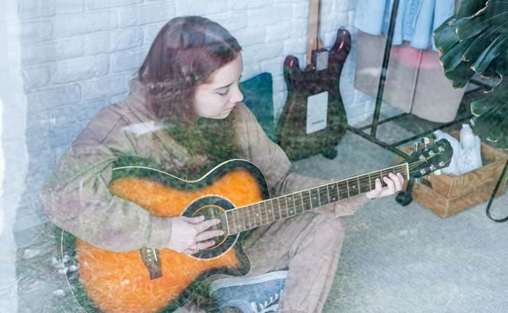 young caucasian red-haired woman sitting on rug and playing acoustic guitar at home, view through the glass window