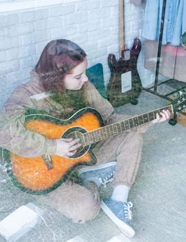 young caucasian red-haired woman sitting on rug and playing acoustic guitar at home, view through the glass window