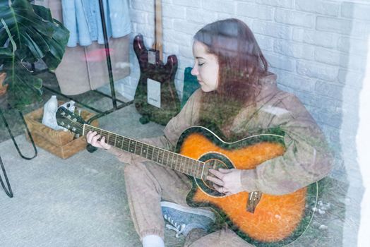 young caucasian red-haired woman sitting on rug and playing acoustic guitar at home, view through the glass window