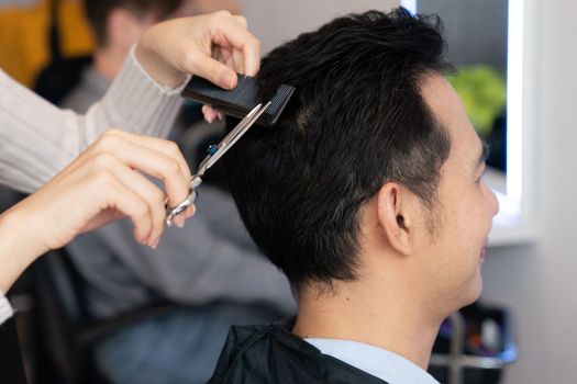 Closeup asian female barber making haircut for male customer with male hairstyle in qualified barbershop. Men's hairstyling by a scissor and comb in hair salon concept.