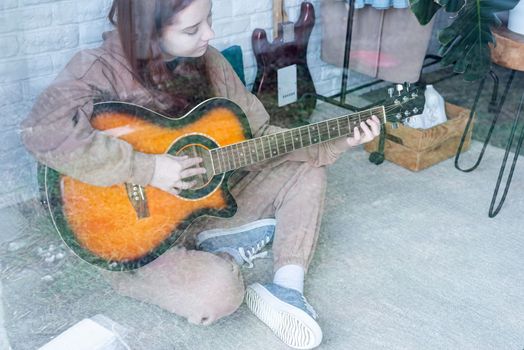 young caucasian red-haired woman sitting on rug and playing acoustic guitar at home, view through the glass window