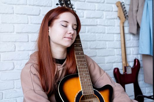 young caucasian red-haired woman sitting on rug and playing acoustic guitar at home