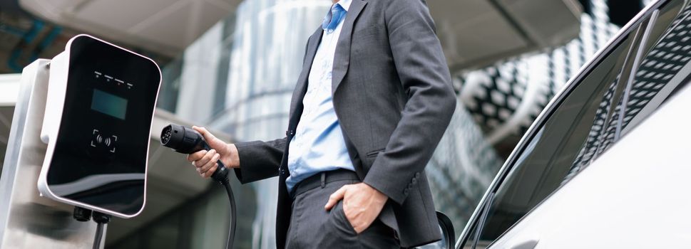 Below view of progressive businessman with electric car recharging at public charging station at modern city residential building. Eco friendly rechargeable EV car powered by alternative clean energy.