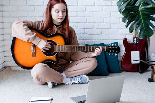young caucasian red-haired woman learning to play acoustic guitar at home, making notes and using laptop