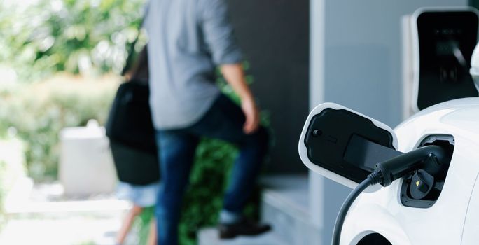 Focus electric car recharging at home charging station with blurred father and daughter walking in background. Progressive green and clean energy vehicle for healthy environment lifestyle concept.