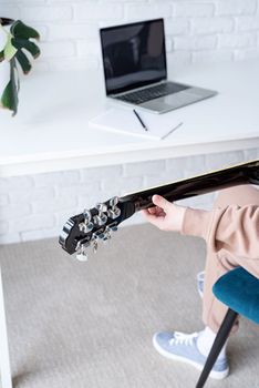 young caucasian red-haired woman learning to play acoustic guitar at home, making notes and using laptop