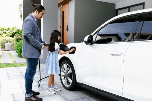 Progressive father and daughter plugs EV charger from home charging station to electric vehicle. Future eco-friendly car with EV cars powered by renewable source of clean energy.