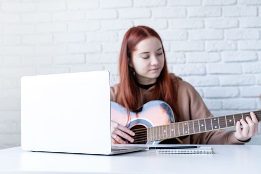 young caucasian red-haired woman learning to play acoustic guitar at home, making notes and using laptop