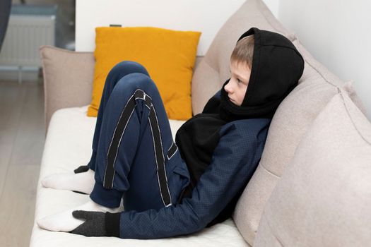 People. Energy crisis concept. A teenage boy sits at home on the sofa in gloves, a warm scarf and a hood on his head, freezes due to the heating being turned off. War in Europe. Cold in apartments. Energy crisis Ukraine. Close-up.