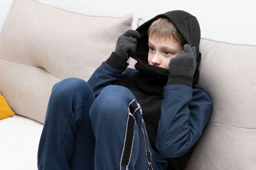 People. Energy crisis concept. A teenage boy sits at home on the sofa in gloves, a warm scarf and a hood on his head, freezes due to the heating being turned off. War in Europe. Cold in apartments. Energy crisis Ukraine. Close-up.