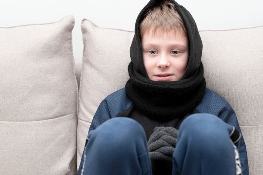 People. Energy crisis concept. A teenage boy sits at home on the sofa in gloves, a warm scarf and a hood on his head, freezes due to the heating being turned off. War in Europe. Cold in apartments. Energy crisis Ukraine. Close-up.