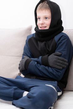People. Energy crisis concept. A teenage boy sits at home on the sofa in gloves, a warm scarf and a hood on his head, freezes due to the heating being turned off. War in Europe. Cold in apartments. Energy crisis Ukraine. Close-up.