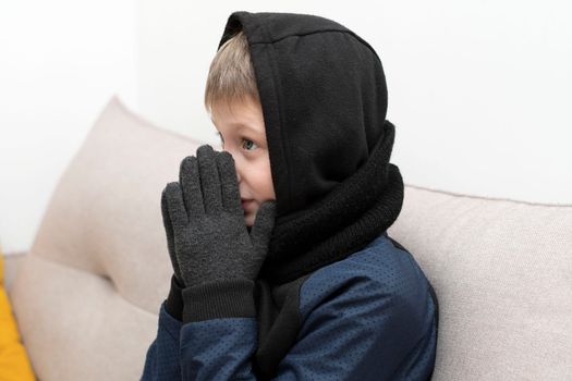People. Energy crisis concept. A teenage boy sits at home on the sofa in gloves, a warm scarf and a hood on his head, freezes due to the heating being turned off. War in Europe. Cold in apartments. Energy crisis Ukraine. Close-up.