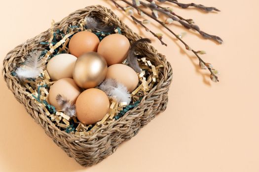 Easter eggs natural and gold in basket with feather. Branch of willow catkins. Light pastel orange background with copy space.