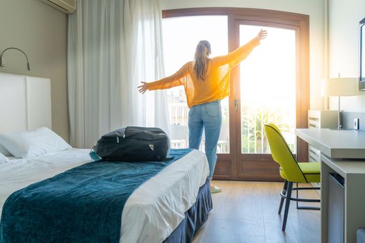 Tourist woman in the hotel bedroom with her luggage raised hands up and standing near the window. Travel and vacation concept. High quality photo