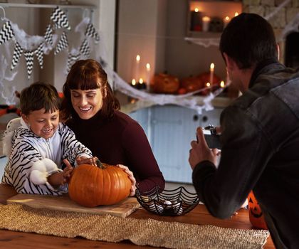 Hes making the scariest looking pumpkin ever. a father taking pictures of his son and wife carving a pumpkin together on halloween at home