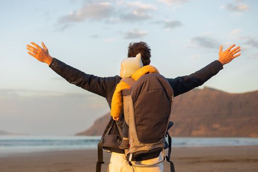 Father rising hands to the sky while enjoying pure nature carrying his infant baby boy son in backpack on windy sandy beach of Famara, Lanzarote island, Spain at sunset. Family travel concept