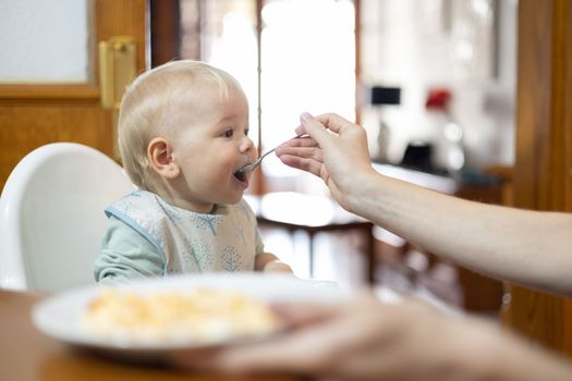 Mother spoon feeding her infant baby boy child sitting in high chair at the dining table in kitchen at home.