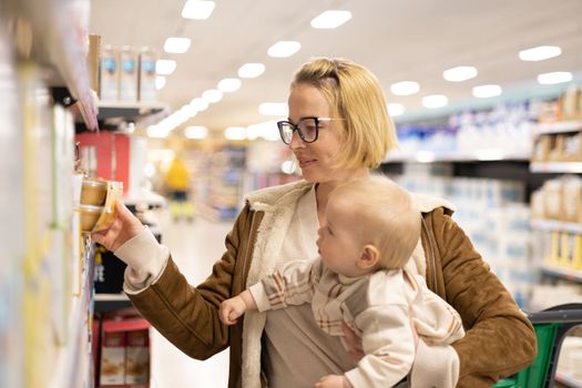 Caucasian mother shopping with her infant baby boy child choosing products in department of supermarket grocery store