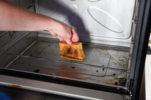 Female hand with sponge and detergent cleaning the kitchen oven from burnt drops, housekeeping concept. Scrubbing the stove, High quality photo