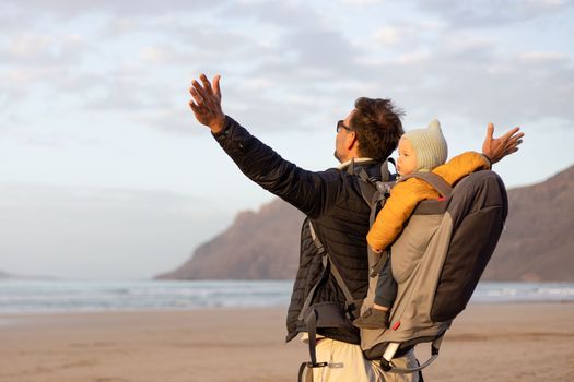 Father rising hands to the sky while enjoying pure nature carrying his infant baby boy son in backpack on windy sandy beach of Famara, Lanzarote island, Spain at sunset. Family travel concept