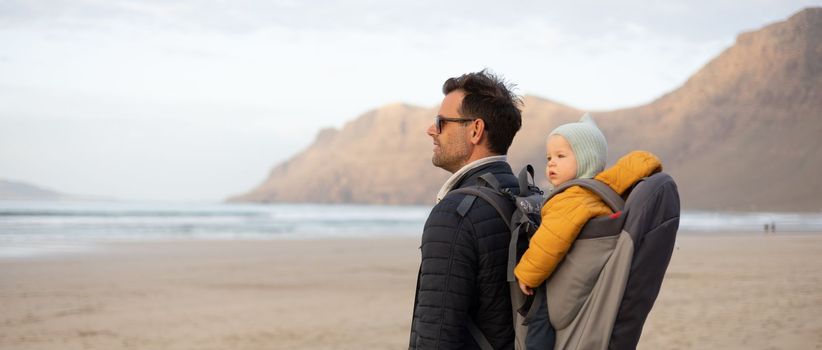 Father rising hands to the sky while enjoying pure nature carrying his infant baby boy son in backpack on windy sandy beach of Famara, Lanzarote island, Spain at sunset. Family travel concept