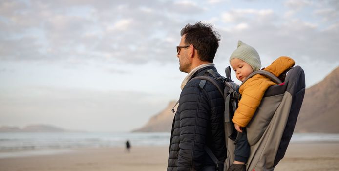 Father rising hands to the sky while enjoying pure nature carrying his infant baby boy son in backpack on windy sandy beach of Famara, Lanzarote island, Spain at sunset. Family travel concept