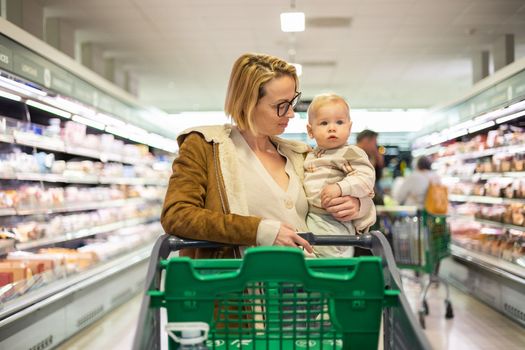 Caucasian mother shopping with her infant baby boy child choosing products in department of supermarket grocery store