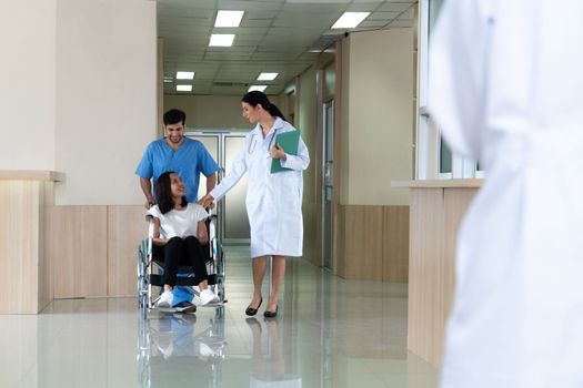 Doctor and male nurse transport a female patient in a wheelchair along sterile hospital corridor. Health care and nursing care for disabled handicapped patient in the hospital concept.
