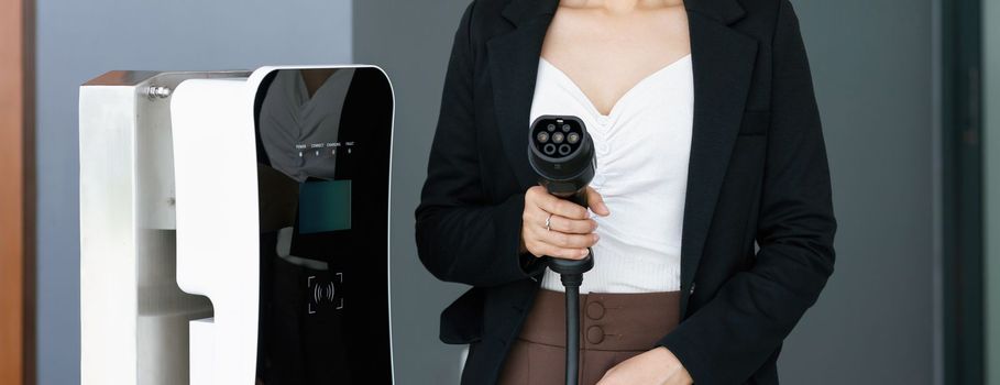 Closeup asian woman holding and pointing an EV plug, a home charging station providing a sustainable power source for electric vehicles. Alternative energy for progressive lifestyle.