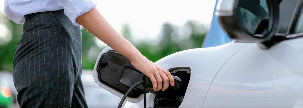 Closeup focus hand insert EV charger to electric vehicle at public charging point in car park with blur business people in backdrop, eco-friendly lifestyle by rechargeable car for progressive concept.