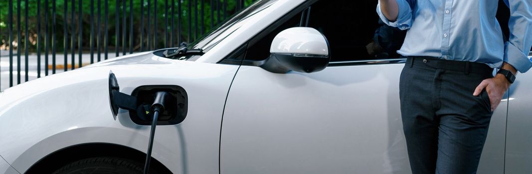 Closeup progressive suit-clad businessman with his electric vehicle recharge his car on public charging station in modern city with power cable plug and renewable energy-powered electric vehicle.