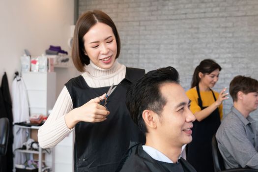 Asian female hairdresser making haircut for two male customer with male hairstyle in qualified barbershop. Men's hairstyling by a scissor and comb in hair salon concept.
