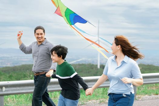 Progressive happy carefree family vacation concept. Young parents mother father and son run along and flying kite together on road with natural scenic on mountain and wind turbine background.