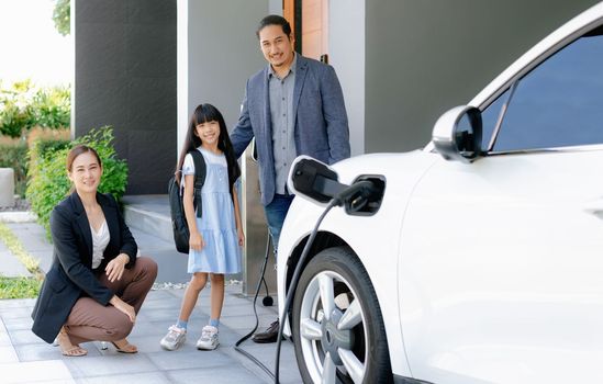 Progressive young parents and daughter with electric vehicle and home charging station. Green and clean energy from electric vehicles for healthy environment. Eco power from renewable source at home.