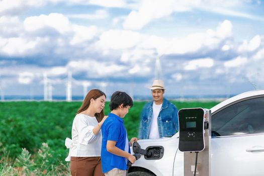 Concept of progressive happy family enjoying their time at wind farm with electric vehicle. Electric vehicle driven by clean renewable energy from wind turbine generator for charging station.