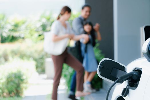 Focus closeup electric vehicle recharging battery from home electric charging station with blurred family in background. Renewable clean energy car for progressive eco awareness lifestyle concept.