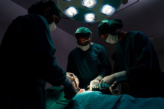 Surgical team performing surgery to patient in sterile operating room. In a surgery room lit by a lamp, a professional and confident surgical team provides medical care to an unconscious patient.