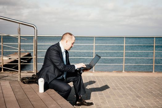 Digital Nomad, a young tattooed man working remotely online, typing on a laptop keyboard while sitting on a beach at sunset. Working remotely on vacation, running an online business from a distance