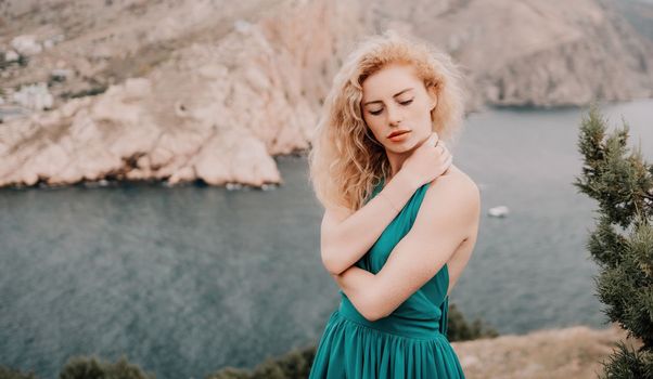 Side view a Young beautiful sensual woman in a mint long dress posing on a volcanic rock high above the sea during sunset. Girl on the nature on overcast sky background. Fashion photo