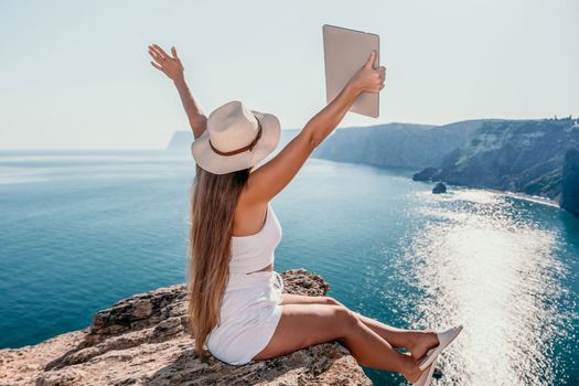 Successful business woman in yellow hat working on laptop by the sea. Pretty lady typing on computer at summer day outdoors. Freelance, travel and holidays concept.