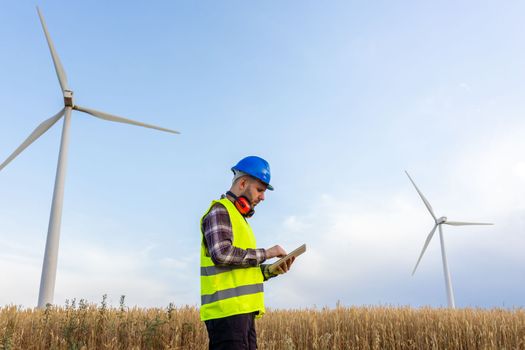 Side view of male maintenance worker wearing helmet and vest checking data on digital tablet in wind turbine farm. Copy space. Renewable energies concept.