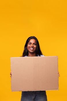Young Indian asian woman looking at camera holding a cardboard banner isolated in yellow background. Studio shot. Vertical image.