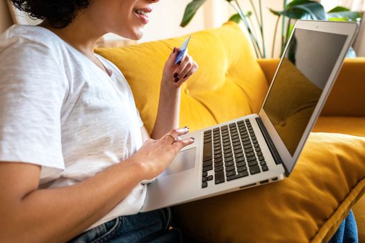 Side view of unrecognizable happy African american woman using credit card and laptop to shop online at home. Lifestyle and e-commerce concept.