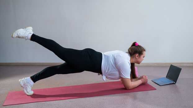 Young fat caucasian woman doing a plank on a sports mat. A cute plus size girl in sportswear is doing fitness exercises and watching an online tutorial on a laptop.