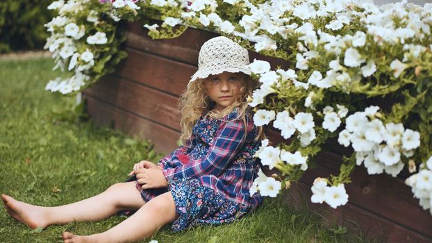 A little girl in a panama hat sits by the white petunias in the garden