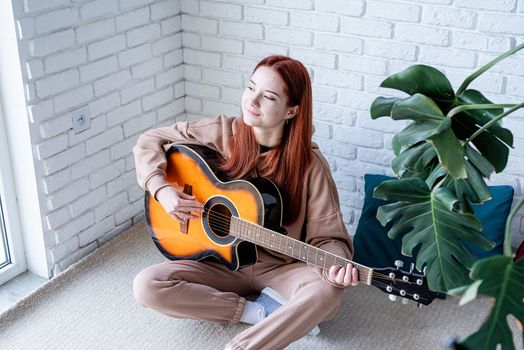 young caucasian red-haired woman sitting on rug and playing acoustic guitar at home