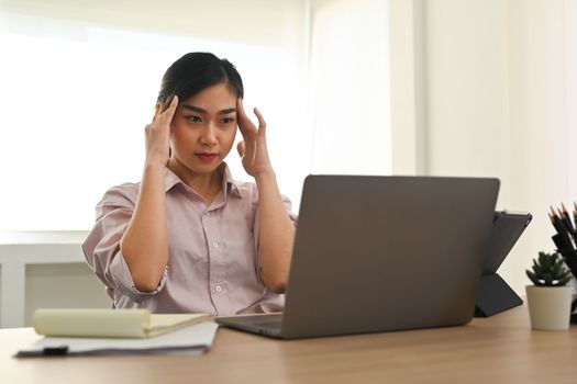 Tired asian woman office worker massaging temples, exhausted from overwork or chronic migraine while sitting at workplace.