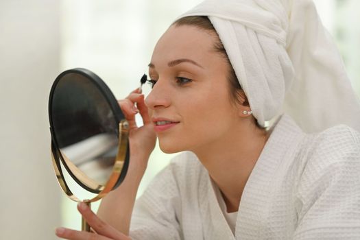 Shot of beautiful caucasian woman n bathrobe applying mascara on eyelashes. Beauty and cosmetics concept.